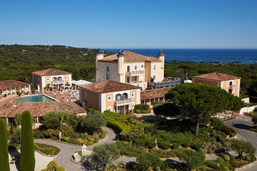 Exterior view of Airelles Château de la Messardière, surrounded by lush trees and a glimpse of the ocean in the background.