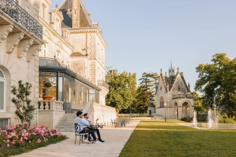 The exterior of Château de Mirambeau is visible as a couple relaxes on chairs in the foreground, enjoying the view.