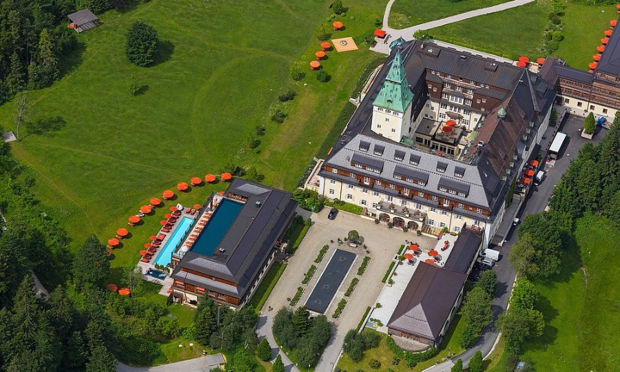 Panoramic aerial image of Schloss Elmau, showcasing a grand castle surrounded by dense forest.